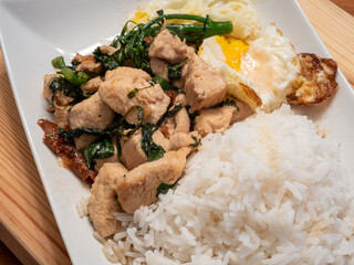Thai basil chicken with fried egg, broccolini and jasmine rice on white plat on top of wooden board.