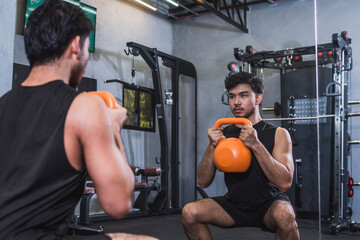 An asian man does a set of goblet squats using an orange kettlebell. Training legs at the gym.