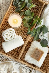 Top-view shot of a woven bamboo tray with rolled towels, a bar of handmade soap, a sprig of eucalyptus, and a flickering candle