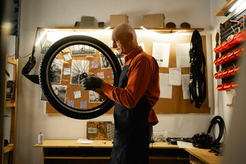 Elderly male worker holding and repairing bicycle wheel while standing in authentic bicycle...