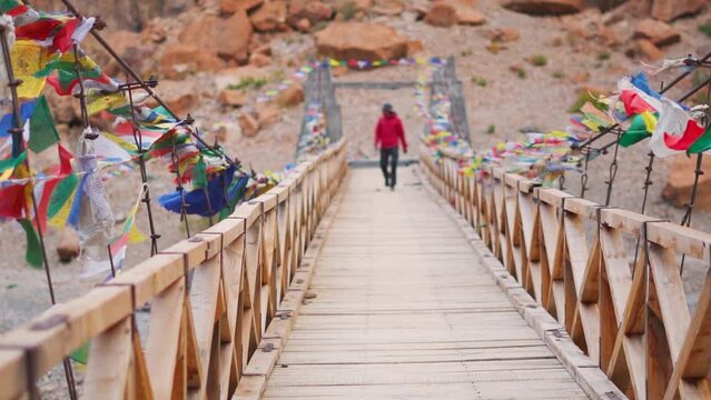 Handheld shot of wooden bridge above the Tsarap Chu river with prayer flags waving on the side due to the strong winds with defocused man walking on the bridge during trek to Phugtal Gompa.	