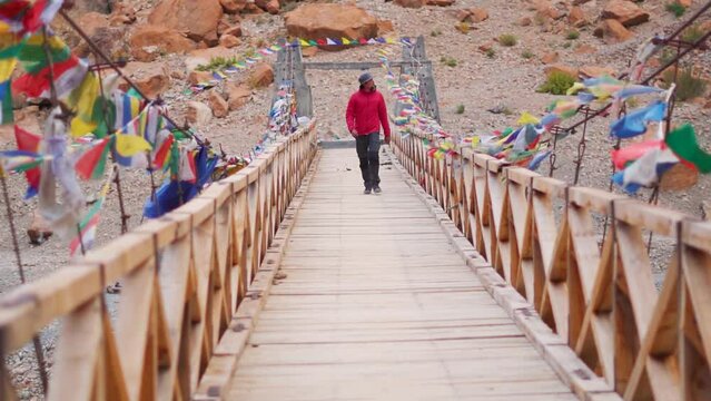 Indian male tourist wearing hat walking on the wooden bridge above the Tsarap Chu river on the way towards Phugtal Monastery at Zanskar Valley in remote regions of Ladakh, India. Tourist on bridge.