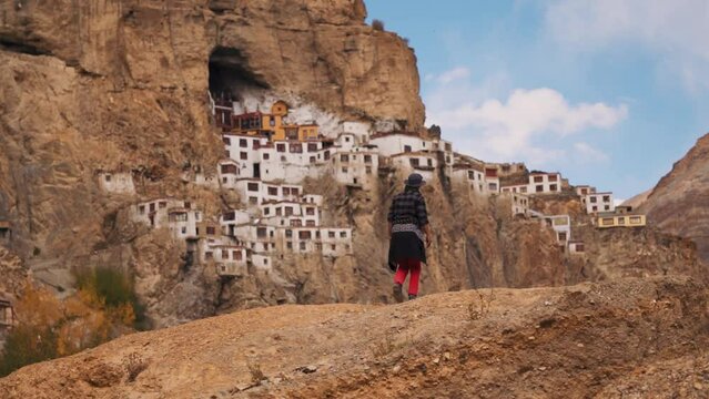 Rear view shot of an Indian male tourist wearing hat walking in front of Old Phugtal Monastery built on a cave in the mountains in the remote regions of Ladakh at Zanskar Valley in India.