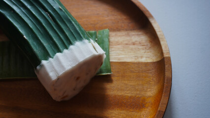 Close up isolated flat lay whole square tempeh wrapped with banana leaf above wooden tray. Selective focus traditional raw side dishes from fermented soybeans           
