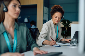Call center agent writing notes while working on laptop in office.