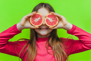 Little cute funny girl uses grapefruit for eyes and has fun in the studio on a green background. Concept of citrus diet, fruits and vitamins.