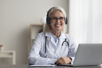 Portrait of mature professional licensed medical worker sit at desk with laptop smile look at...