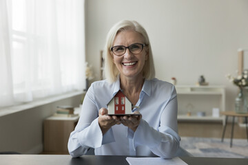 Portrait of happy mature Caucasian woman realtor holding small miniature of modern house, smile, look at camera. Provide professional consultation, selling real-estate property, remodelling services