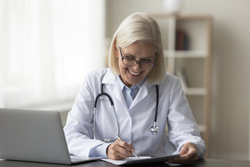 Mature woman doctor sit at desk make notes, jotting information, prepare paper document, reviewing medical records, completing administrative tasks, filling forms or writing prescriptions at workplace - obrazy, fototapety, plakaty