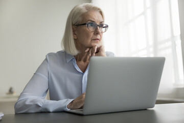 Pensive older businesswoman sit at desk, working on laptop, think over marketing report at workplace, feel doubt, analyze agreement terms, make decision, ponder task or strategy. Brainwork, technology