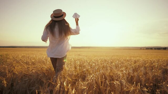 Carefree woman running on wheat field throwing paper plane in sky keeping straw hat on head at sunset, back view. Freedom, farm lifestyle, resting in countryside lifestyle, outdoor activities concept.