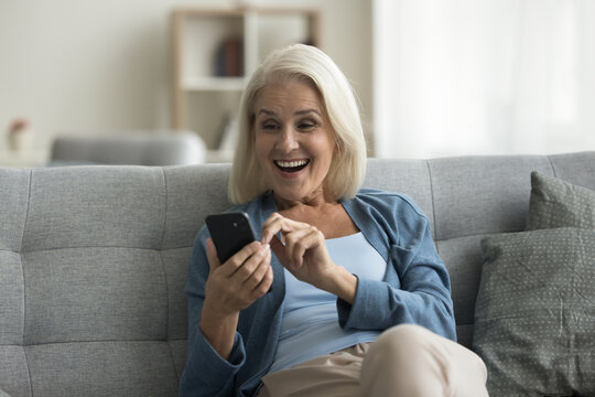 Overjoyed mature woman sit on sofa gawp at cellphone screen with mouth opened, looks amazed, feel surprised, read unbelievable great news about lottery win, huge discount, sell-out commercial offer