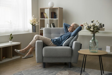 Smiling attractive senior woman relax at home, enjoy lazy weekend seated on armchair with hands...