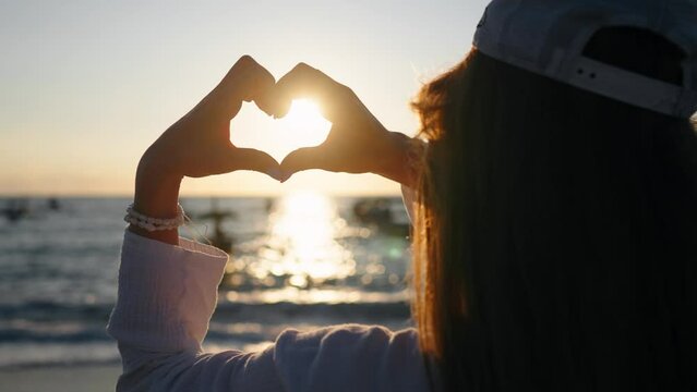 Woman showing heart symbol on ocean. Young lady doing heart gesture with hands making heart shape at seaside, admire sea sunset. Love, romantic, freedom, relaxation, calm, summer vacation concept.