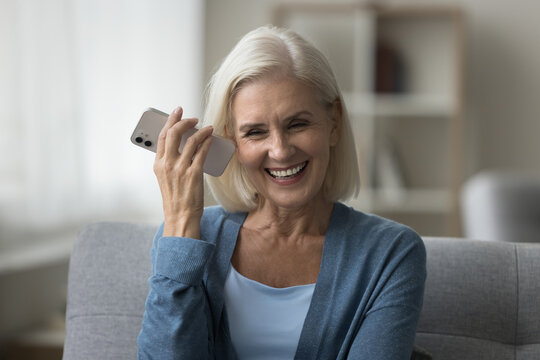 Smiling middle-aged woman sit on sofa at home, holding mobile phone near ear, listening audio message, chatting with relatives or friend, sharing voicemail, enjoy remote communication on speakerphone