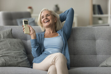 Happy peaceful middle-aged woman holding remote controller, turning switch-on air conditioner...