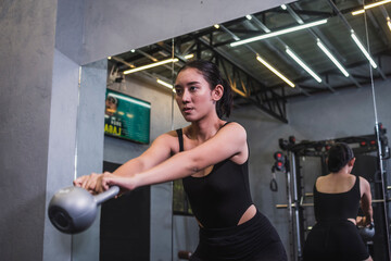 A young asian woman vigorously does a set of kettlebell swings. Doing cardiovascular exercise, weigh training or High Intensity Interval Training. Wearing a bodysuit and shorts.