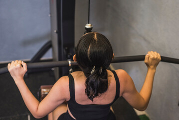 A young asian woman performs wide grip straight lat pulldowns. Weight training targeting back muscles and upper body. Shot from behind. - obrazy, fototapety, plakaty