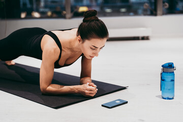 Beautiful girl training in the gym. Young woman performs an abdominal plank while checking the run...
