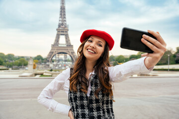 Beautiful young woman visiting paris and the eiffel tower. Parisian girl with red hat and fashionable clothes taking a pov selfie with the smartphone camera and smiling. - obrazy, fototapety, plakaty