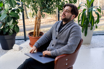 of young guy office worker against the background of a window sitting on a chair working at a laptop