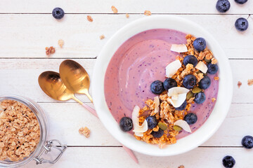 Healthy blueberry and coconut smoothie bowl with granola. Overhead view table scene on a white wood background.