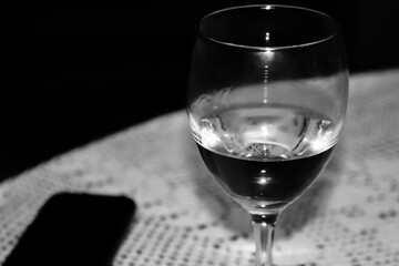 A quarter filled wine glass with a cell phone on the side on a table covered with white embroidered tablecloth. Black and white photo