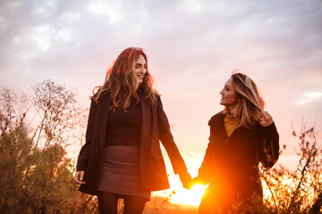 Two happy women girlfriends holding hands walking through autumn field at sunset in countryside