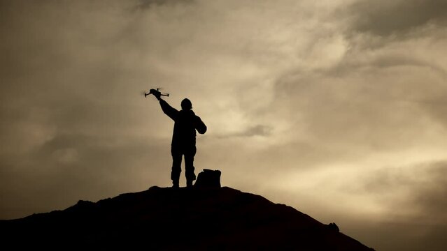 A solitary soldier's silhouette stands out on a hilltop as he launches a drone into the twilight sky, with the dramatic backdrop of a vibrant sunset providing a powerful contrast. Camera 8K RAW.