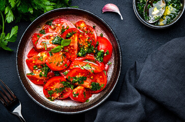 Simple summer juicy tomato salad with parsley, dill, garlic and olive oil dressing, black stone table background, top view