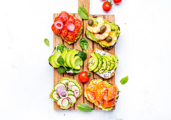 Avocado toasts with salmon, shrimp, vegetables, spinach, capers and cream cheese, served on wooden board, white table background, top view - 783906451