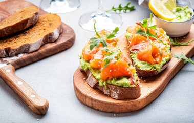 Avocado sandwich with salmon on rye bread with guacamole sauce, arugula and sesame seeds on wooden board, rose wine glass on gray table background, top view - 783906878