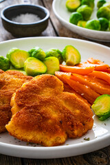 Crispy breaded fried pork loin chops with sweet potato fries and boiled Brussels sprouts on wooden table
