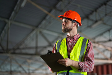 A man wearing a yellow vest and an orange hard hat is holding a clipboard. He is looking up at...
