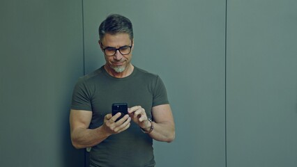 Casual, mature man in glasses focused on texting on phone with a plain gray background, reading emails, porting to social media, using app.
