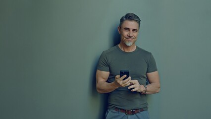 Portrait of a stylish middle-aged man smiling and holding a smartphone. Happy, confident mid adult male in casual. Blank copy space on a gray wall background.
