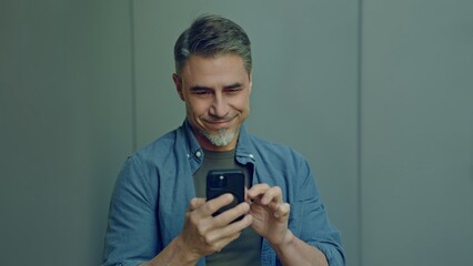 Portrait of a content middle-aged man looking at cell phone. Happy, confident mid adult male in casual. Blank copy space on a gray wall background.