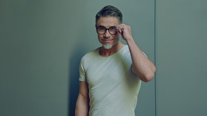 Portrait of a stylish middle-aged man adjusting glasses and smiling. Happy, confident mid adult male in casual. Blank copy space on a gray wall background.