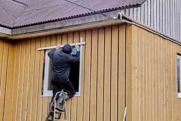 Standing on steel folding stepladder, construction worker installs external slopes on window of rural house from facade. - obrazy, fototapety, plakaty