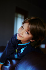 Smiling little boy leans against back of leather sofa in dark room.