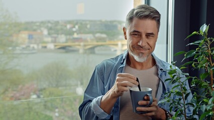 Casual older man standing by window at at home drinking morning coffee. Portrait of happy mid adult, middle aged man in 50s, smiling. Cityscape with river and bridge in background. - 783940694