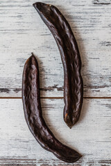 Two dried carob pods parallel to each other on a textured white wooden background. - obrazy, fototapety, plakaty