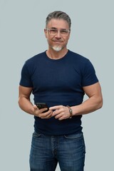 Portrait of mid adult, older, gray hair man in glasses holding phone. Male in casual and jeans standing isolated on gray background. Happy confident smile.