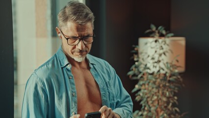 Mid adult man standing in sunlight in open shirt in the morning, checking email, news or social media on cell phone. Happy older male smiling in glasses.