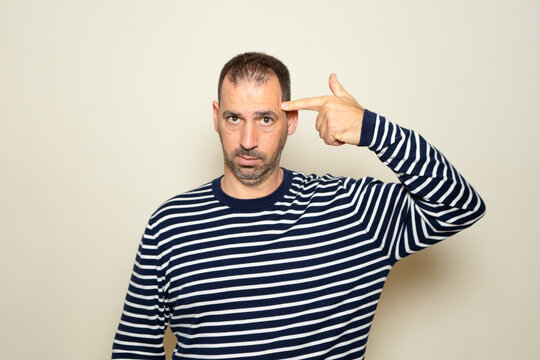 Hispanic man with beard in his 40s wearing a striped sweater standing over a beige background shooting and killing himself pointing with hand and fingers at his head like a gun, suicidal gesture