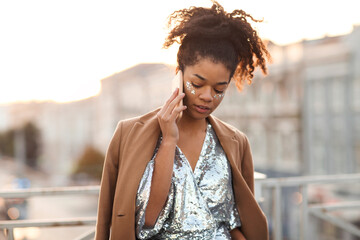 African american woman with glitter on cheekbones in sequin dress talking on mobile phone at rooftop