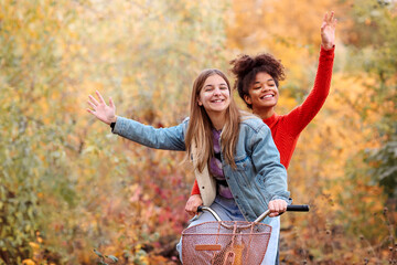 Two young happy diverse female best friends riding bicycle together in autumn forest, active positive multiracial female friends
