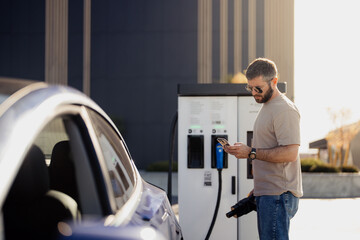 A man uses his smartphone while charging his electric car at a charging station. Modern, urban...