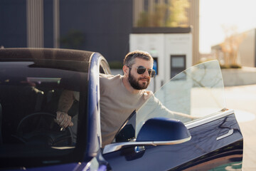Stylish bearded man in sunglasses getting out of a modern blue electric car, showcasing eco-friendly transportation.
