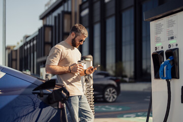 A man checks his smartphone at a charging station while his electric vehicle charges. The scene...